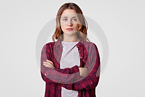 Indoor shot of serious self assured teenage girl keeps hands crossed, wears checkered shirt, looks seriously at camera, isolated