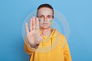 Indoor shot of serious woman in yellow hoodie with Ukrainian flag on cheeks, showing stop gesture with her palm, not war, peace in