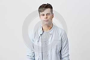 Indoor shot of serious young european male frowns face and pouts lips, being offended by friend who jokes at him