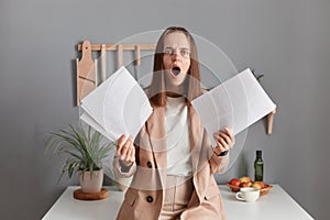Indoor shot of shocked amazed surprised young adult woman wearing beige suit, holding paper documents, having troubles with