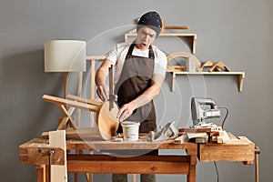 Indoor shot of shocked amazed young adult man carpenter wearing white t-shirt, black cap and brown apron working, joiner at his