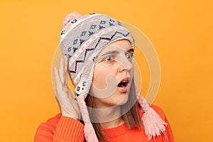 Indoor shot of shocked astonished woman wearing orange sweater and knitted earflap hat standing isolated over yellow background,