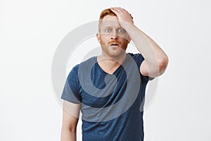 Indoor shot of shocked and stunned male with red hair and bristle, punching himself at forehead and staring shook at