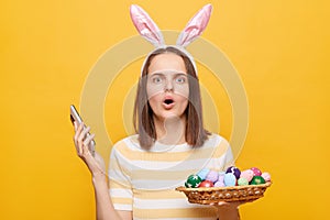 Indoor shot of shocked woman wearing rabbit ears holding Easter eggs in basket and cell phone, looking at camera with big eyes and