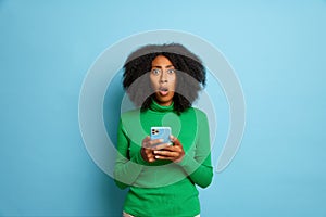 Indoor shot of shocked young woman with curly hair uses mobile phone for online shopping, wears green turtleneck, poses
