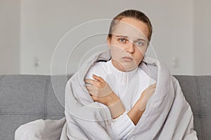 Indoor shot of sick unhappy woman being wrapped in plaid, looking at camera with sad expression, suffering influenza or grippe photo