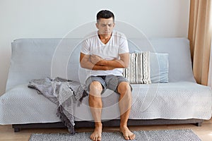 Indoor shot of sick unhealthy man wearing white T-shirt sitting on sofa in living room, holding his belly, suffering stomach ache