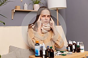 Indoor shot of sick unhealthy young adult woman wearing warm jumper and scarf sitting on sofa in living room at home, massaging