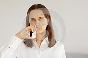 Indoor shot of sick winsome female with pleasant appearance wearing white casual style shirt, using nasal spray, suffering runny