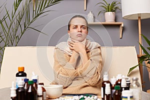 Indoor shot of sick young adult woman wearing warm jumper and scarf sitting on sofa in living room at home, holding her neck,