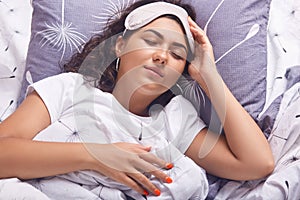 Indoor shot of sleeping girl with blindfold on her head, female wearing white t shirt, lying in bed under blanket, keeps eyes