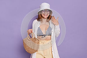 Indoor shot of smiling cheerful woman wearing panama, white shirt and swimming suit, holding bag, expressing pleasant surprise,