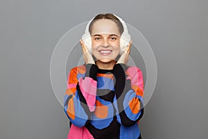 Indoor shot of smiling delighted young adult woman with brown hair wearing jumper and warm earmuffs standing  over gray