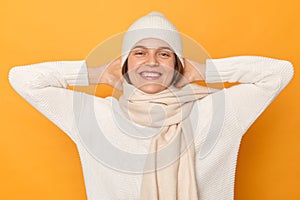 Indoor shot of smiling delighted young adult woman wearing warm sweater, cap and scarf posing isolated over yellow background,