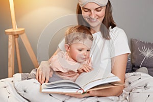 Indoor shot of smiling european woman wearing white t-shirt and blindfold sitting in bed with her toddler daughter, mommy reading