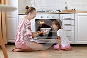 Indoor shot of smiling happy family, mother and daughter sitting on floor near oven and smelling tasty dessert, expressing