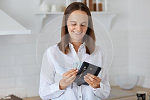 Indoor shot of smiling happy female wearing white shirt standing at home in kitchen with wallet full of money in hands, planning