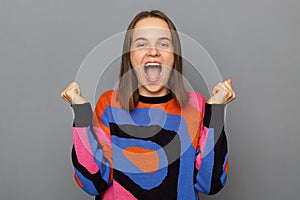 Indoor shot of smiling joyful cheerful woman with brown hair wearing colorful jumper posing isolated over gray background,