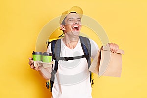 Indoor shot of smiling laughing funny deliveryman with thermo backpack in white T-shirt and cap standing against yellow wall,