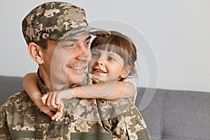 Indoor shot of smiling man soldier wearing camouflage uniform and cap, posing with his daughter, military guy looking at his