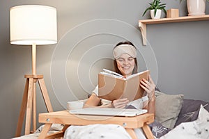 Indoor shot of smiling positive woman wearing white t-shirt and blindfold sitting in bed in front of folded laptop and reading