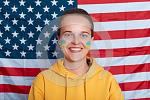 Indoor shot of smiling satisfied woman wearing yellow hoodie with Ukrainian flag on cheeks, being grateful for support from USA,