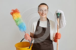 Indoor shot of smiling woman housekeeper wearing brown apron and white t shirt cleaning her apartment, holding mop, ppduster and