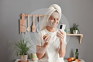 Indoor shot of smiling woman with treatment facial mask enjoying spa procedures at home, posing in the kitchen, eating apple and