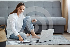 Indoor shot of smiling woman wearing white shirt and jeans sitting on floor near sofa, using laptop, holding credit card, checking