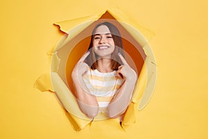 Indoor shot of smiling young adult woman stands in torn paper hole, pointing at her perfect smile and teeth, looking through