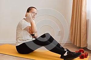 Indoor shot of sportswoman touching painful hand, sitting on mat, suffering joint injury after yoga, sport trauma elbow