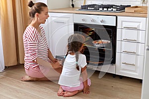 Indoor shot of surprised amazed woman wearing striped shirt and child in white t shirt sitting on floor near oven and looking