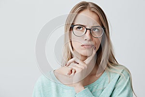 Indoor shot of thoughtful pretty woman has long blonde hair with stylish eyewear, looks aside with pensive expression