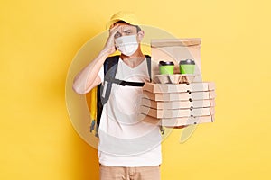 Indoor shot of tired deliveryman in protective mask standing against yellow wall, holding pizza and coffee, suffering headache,