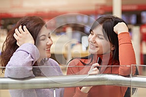 Indoor shot of two woman talking beside clothes rail in shopping mall, look happy, females gossiping, ladies wearing casual