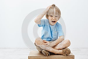 Indoor shot of unhappy cute blond child with vitiligo, having two-colored skin, sitting on floor with crossed feet