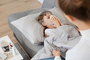 Indoor shot of unhealthy ill female child with handkerchief lying on sofa under blanket suffering runny nose and high temperature