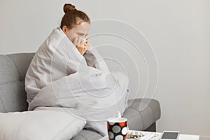 Indoor shot of unhealthy woman with hair bun sitting on sofa wrapped in blanket, catching cold in autumn in cold weather,