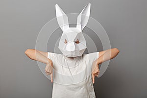 Indoor shot of unrecognizable woman wearing white t shirt and paper rabbit mask standing isolated over gray background, pointing