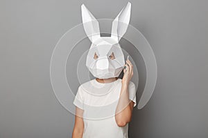 Indoor shot of unrecognizable woman wearing white t shirt and paper rabbit mask talking on cell phone, looking at camera, standing