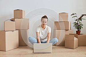 Indoor shot of woman freelancer with bun hairstyle wearing white T-shirt sitting on the floor near cardboard boxes with stuff and