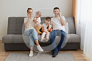 Indoor shot of woman and man sitting on couch and talking phones with their friends, wearing casual style clothing, posing with