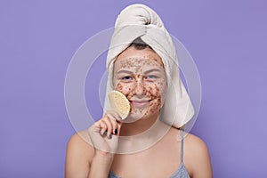 Indoor shot of woman with sponge in handnear her face, lady needs to remove facial mask off, posing isolated over lilac background