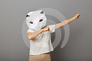 Indoor shot of woman wearing cat paper mask and white T-shirt posing isolated over gray background, raised hands in dab dance pose