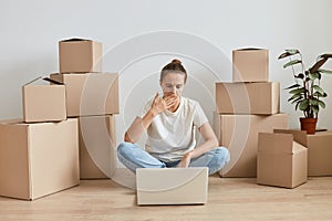 Indoor shot of woman wearing white t shirt sitting on floor surrounded with cardboard boxes with belongings and working on laptop