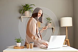 Indoor shot of young adult adorable happy businesswoman wearing jacket working on home office and using laptop and mobile phone,