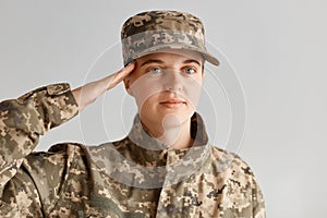 Indoor shot of young adult attractive woman army soldier saluting, looking at camera with serious facial expression, wearing