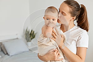 Indoor shot of young adult attractive woman holding daughter wrapped in towel after bathroom, mother kissing her lovely baby girl