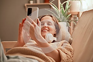 Indoor shot of young adult attractive woman wearing headphones using smart phone while lying on sofa, smiling happily, enjoying