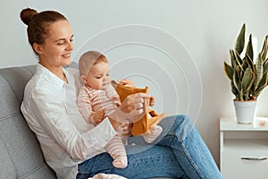 Indoor shot of young adult Caucasian mother sitting on sofa with her infant daughter and holding child`s clothing, having positiv